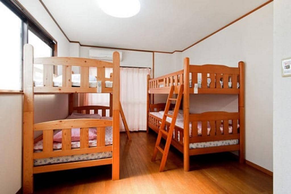 The bright dormitory room features two sets of sturdy wooden bunk beds, accommodating multiple guests. Soft bedding in warm colors is visible on each mattress. Natural light enters through a large window, and a curtain separates the sleeping area from the adjoining space.