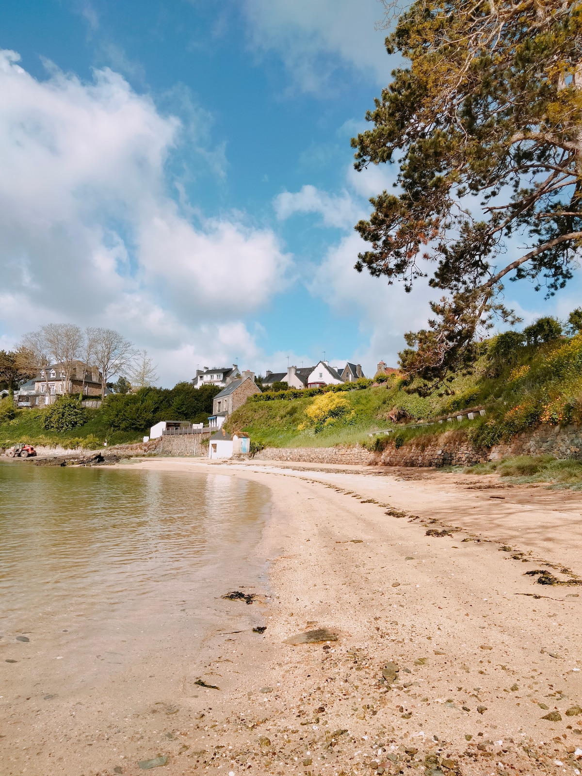 A sandy beach curves along calm waters, leading to a grassy hillside dotted with trees and buildings in the distance. The shoreline displays gentle waves lapping at the sand, while scattered clouds enhance the blue sky above.