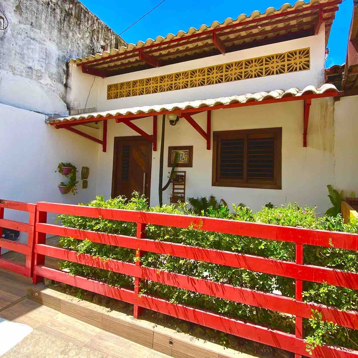 A charming entrance features a red railing that surrounds a lush green garden. The white façade of the building is complemented by wooden windows and a decorative, patterned frieze above. The sunny blue sky adds brightness to the welcoming atmosphere.