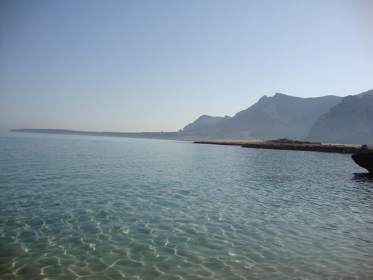 Calm waters reflect the clear sky, with distant mountains framing the coastline. Gentle ripples create patterns on the surface, leading to a rocky shoreline that extends towards the horizon. The serene atmosphere and natural beauty invite relaxation and exploration.