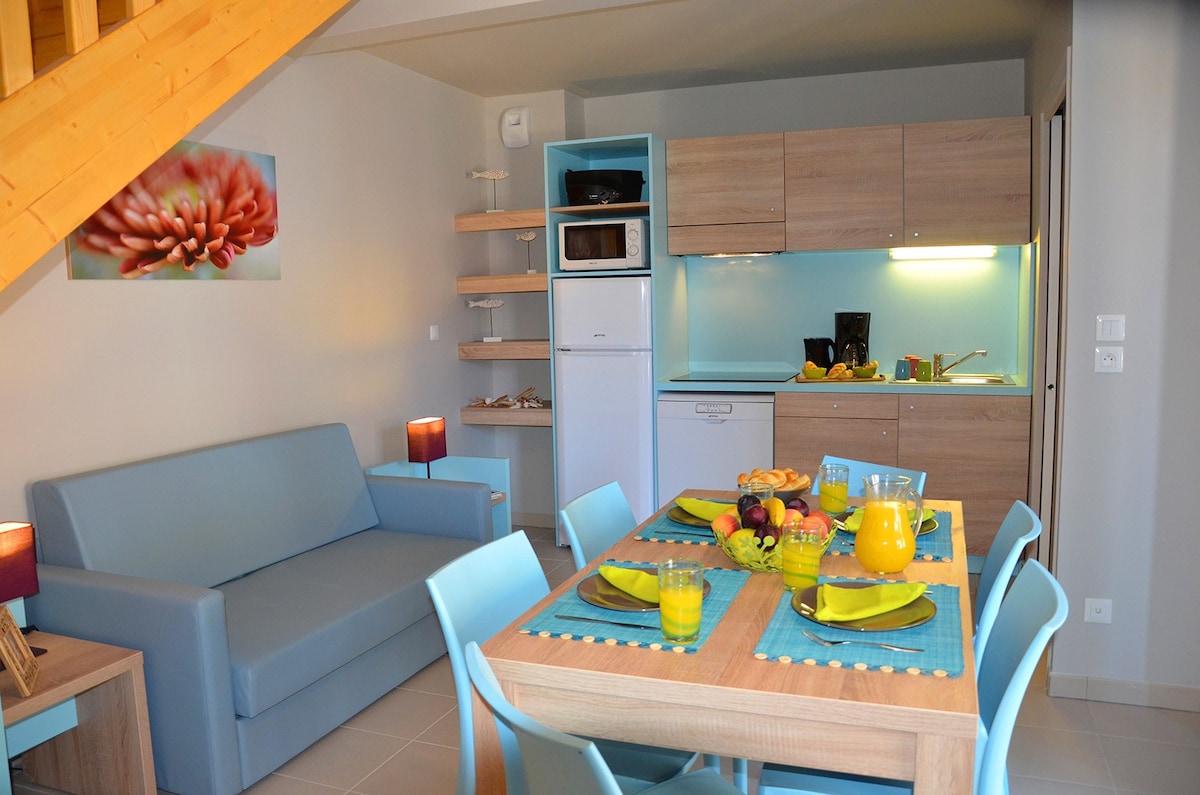 The living space features a modern kitchenette with light wood cabinetry and stainless-steel appliances. A dining table is set with colorful dishes and fresh fruit, complemented by a cozy sofa. Soft lighting from lamps enhances the cheerful and organized environment.