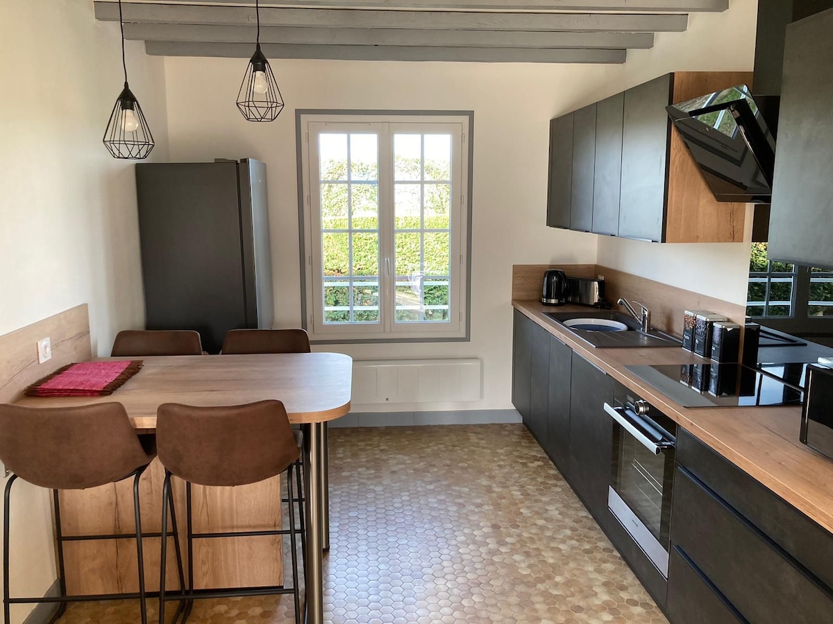 A modern kitchen space features sleek cabinetry with wooden accents and a large window allowing natural light. A dining table with four high-backed chairs sits adjacent to the counter, complemented by minimalist pendant lighting. Appliances include a refrigerator, kettle, and a built-in oven.