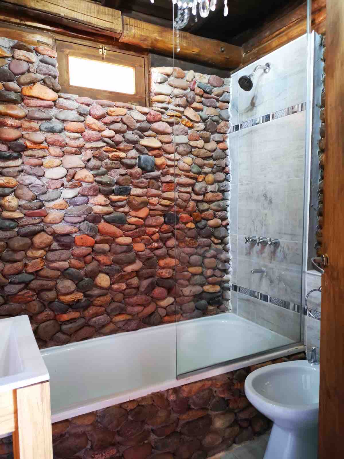 A bathroom showcases a blend of natural stone walls and modern fixtures. A bathtub is positioned next to a glass shower enclosure, and a small window provides natural light. A toilet completes the space, emphasizing a functional and clean design.
