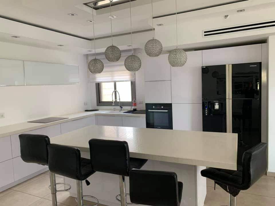 A modern kitchen features sleek white cabinetry and a spacious island with four black bar stools. The stainless steel appliances include a refrigerator and an oven, with textured pendant lights hanging above the island, illuminating the open space.