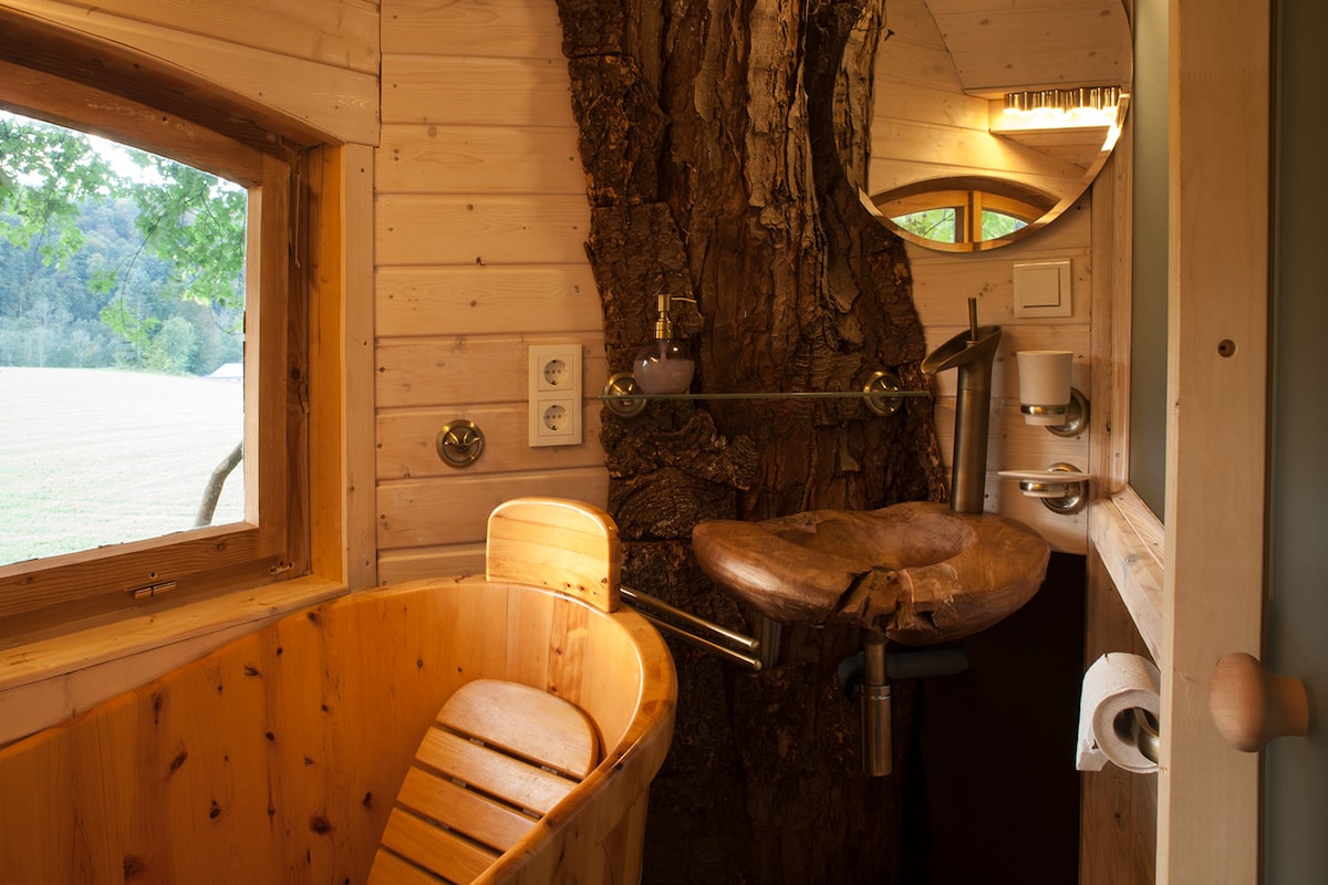 An interior view of the bathroom showcases a wooden bathtub crafted from natural materials. A unique teak wood sink is positioned adjacent to the window, offering a view of the outside landscape. Soft lighting highlights the wood paneling and circular mirror above the sink.