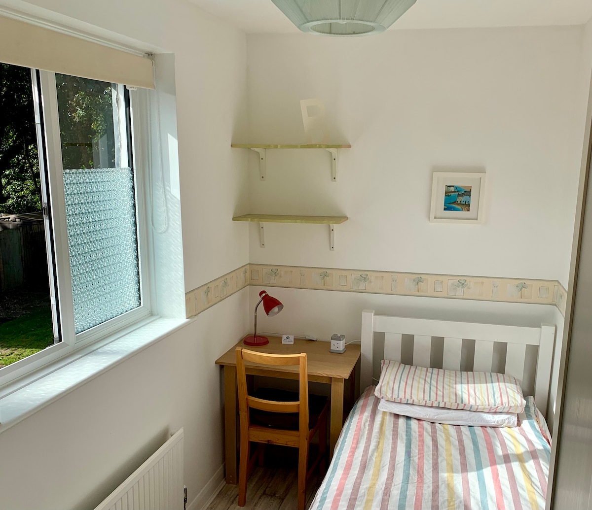 A bright room is showcased, featuring a single bed with striped bedding. A wooden desk and chair are positioned next to the bed, with a desk lamp providing light. Shelving is mounted on the wall above, while a window allows natural light to enter.