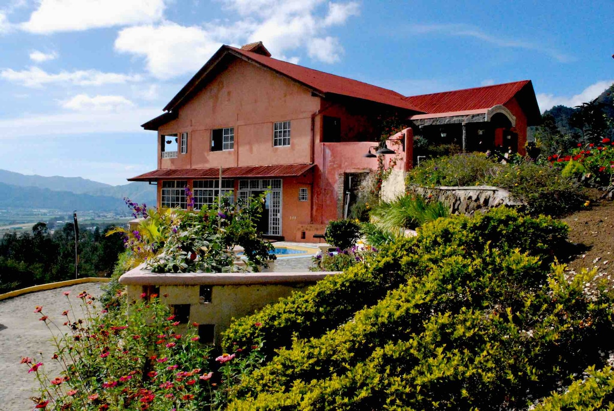 A vibrant pink house with a red roof is set against a mountainous backdrop, surrounded by lush greenery and colorful flower beds. A jacuzzi is located at the forefront, inviting relaxation with scenic valley views in the background under a clear blue sky.