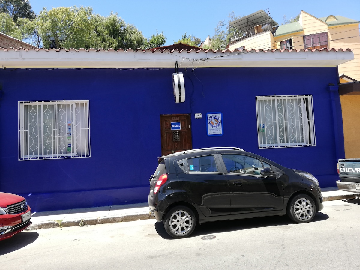 A vibrant blue exterior is showcased on a single-story building, featuring white window bars and a wooden front door. Parked vehicles line the street, with a black car near the foreground, contributing to a lively urban atmosphere.