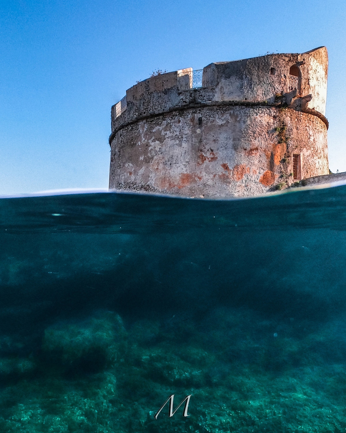 The historic Torre di Capo Galera is captured from a unique underwater perspective, revealing its partially eroded stone structure above the clear blue water. Beneath the surface, the rocky seabed can be seen, surrounded by serene, transparent waves.