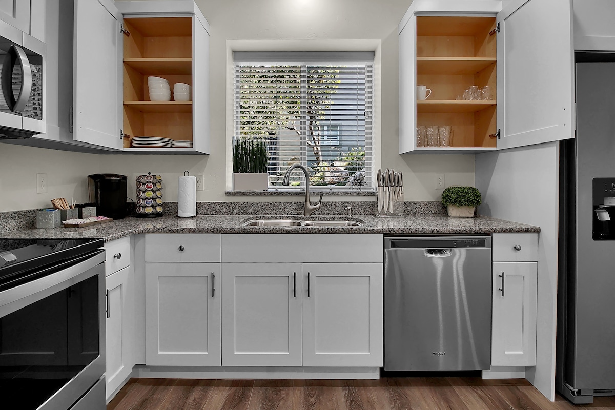 A modern kitchen features sleek cabinetry and a spacious countertop. Stainless steel appliances, including a dishwasher, are present. Natural light enters through a window, illuminating the organized shelves above the sink. Essentials such as a coffee maker and neatly arranged dishware are visible.