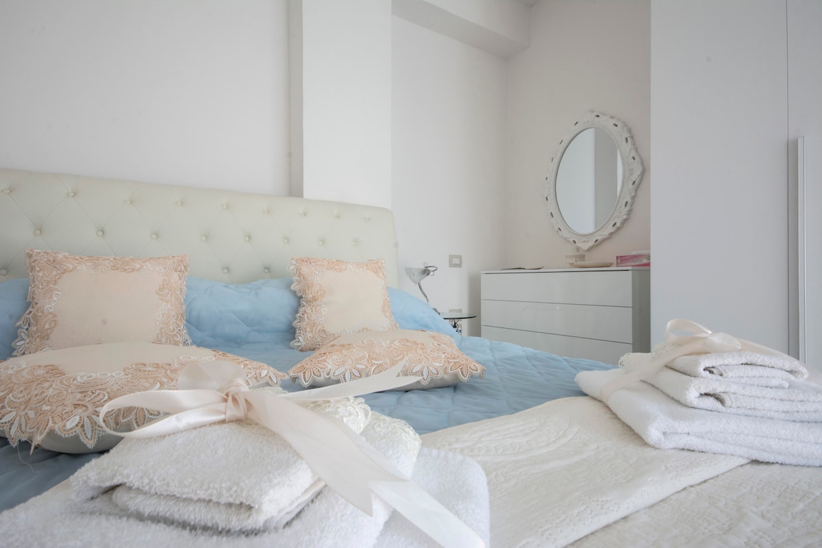 A spacious bed is adorned with decorative pillows, and light blue bedding is neatly arranged. Fresh towels are stacked on the bed, alongside a soft blanket. A round mirror and a dresser are visible in the softly lit room, enhancing the serene atmosphere.