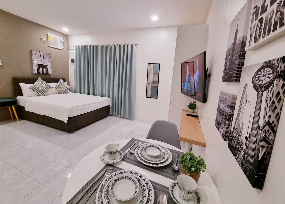 A studio room is depicted with a spacious bed draped in white linens, complemented by decorative pillows. A small dining area with a round table is set with dishware, while a television displays programming. Floor-to-ceiling curtains and artwork enhance the modern decor.