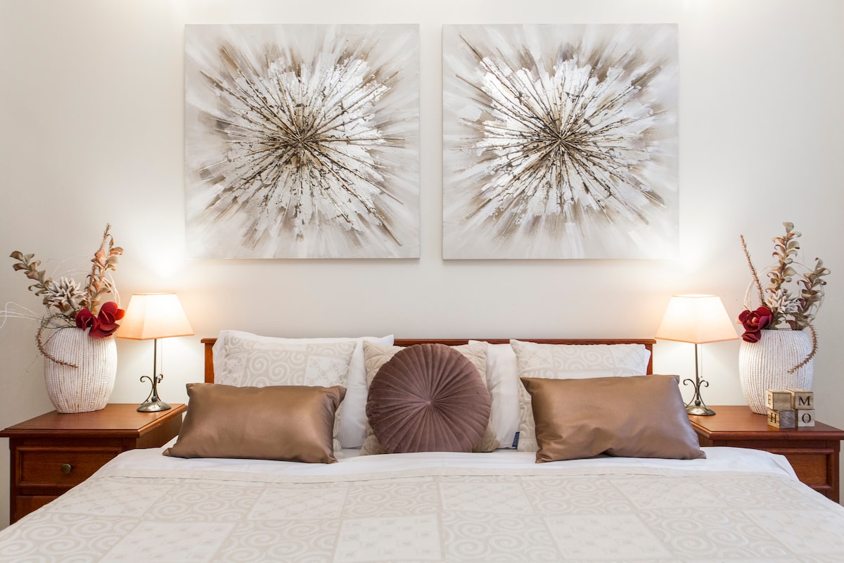 The bedroom features a spacious bed adorned with neutral-toned linens and decorative pillows. Two elegant lamps provide soft lighting on either side of the bed, complemented by large art pieces on the wall above. Decorative vases with dried flowers add a touch of elegance to the room.