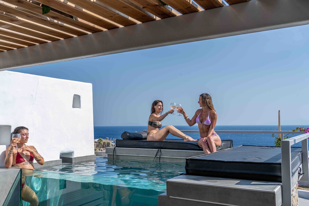 A private pool area features a sleek glass pool with loungers, overlooking the sea. Two guests are seated on the loungers, raising glasses in a toast. The minimalist design and sunny sky create an inviting space for relaxation.