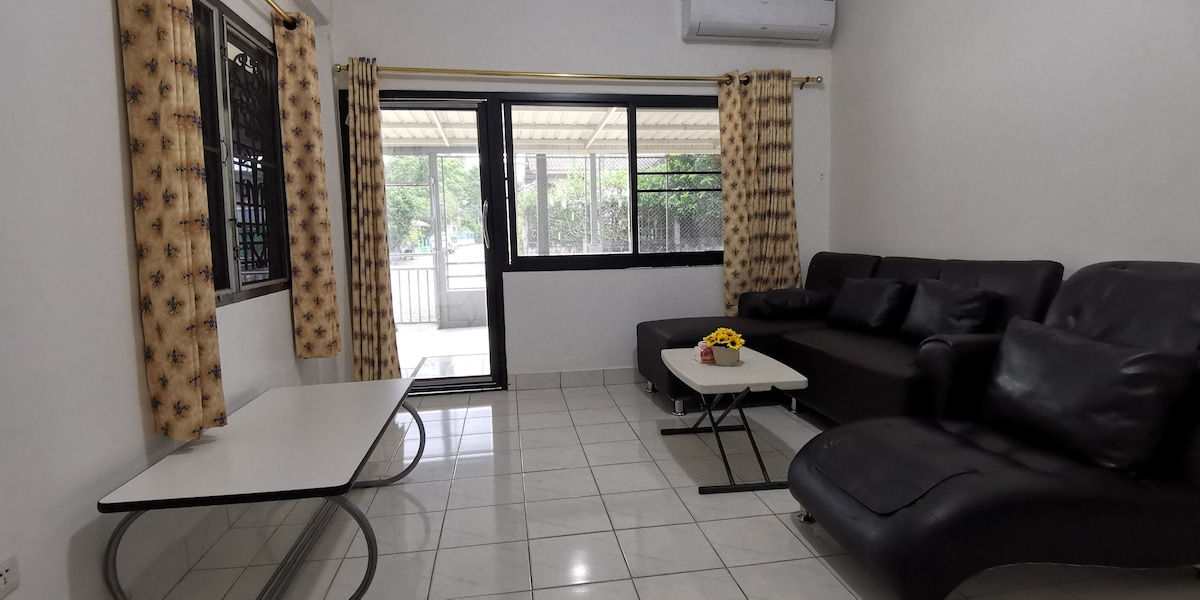 An inviting living area is showcased, featuring a dark sectional sofa and a stylish lounge chair. A coffee table sits in front of the sofa, while large glass doors offer a view of the outdoors. Light curtains frame the windows, enhancing the bright ambiance.