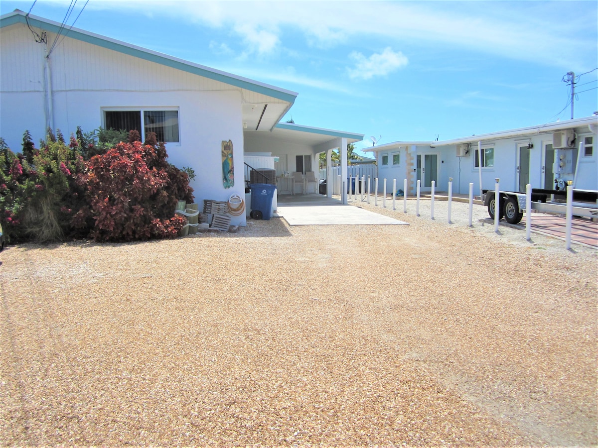 A wide, gravel driveway is seen leading to the duplex building. Colorful foliage borders the space, and utility poles are visible in the background. The parking area accommodates vehicles comfortably, with additional spaces towards the right for guests or boats.