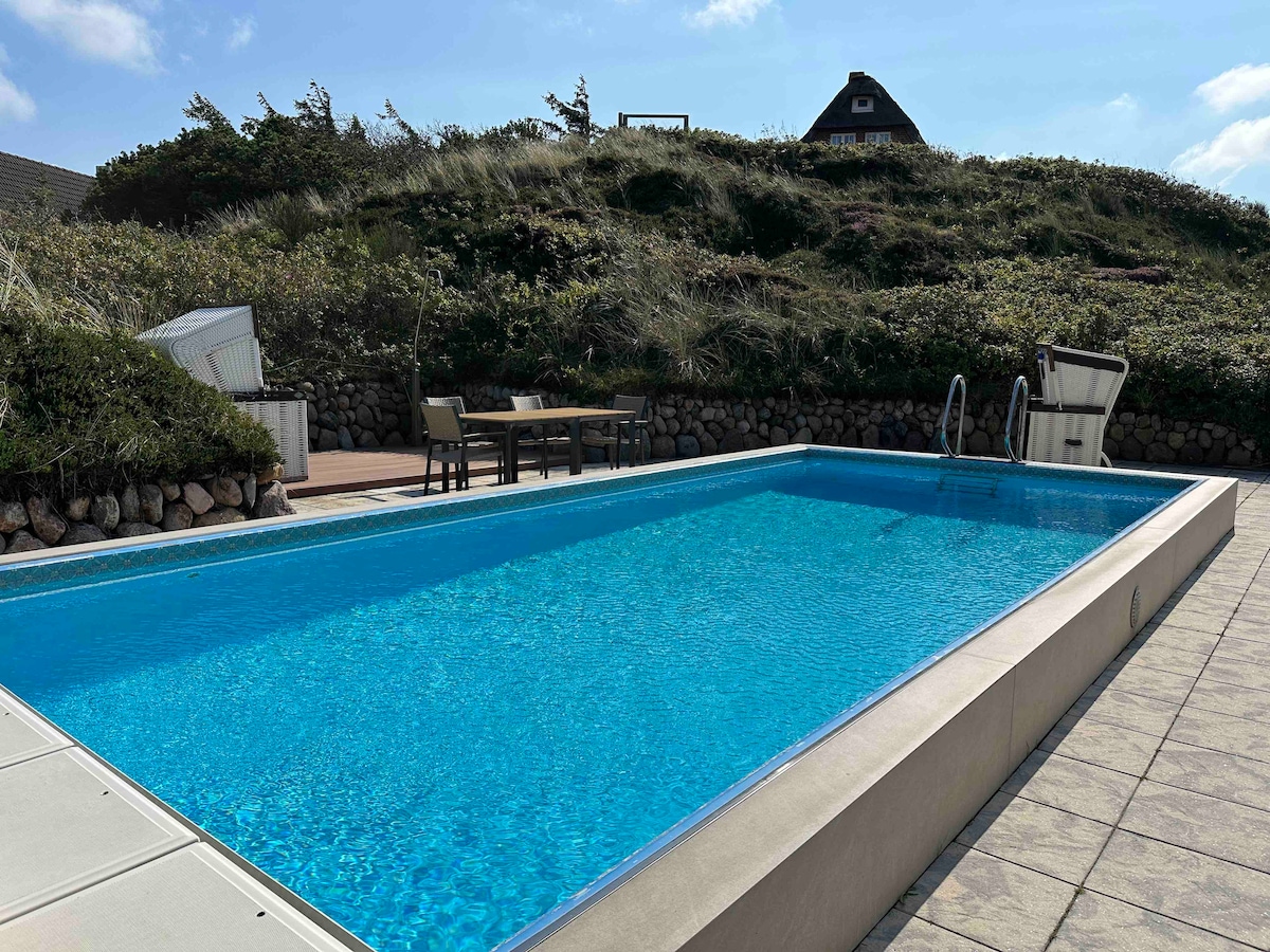 A large outdoor heated pool is surrounded by a patio area featuring lounge chairs and dining furniture. Lush greenery and dunes rise in the background, with a clear blue sky overhead. The pool's crystal-clear water captures reflections of the surroundings.