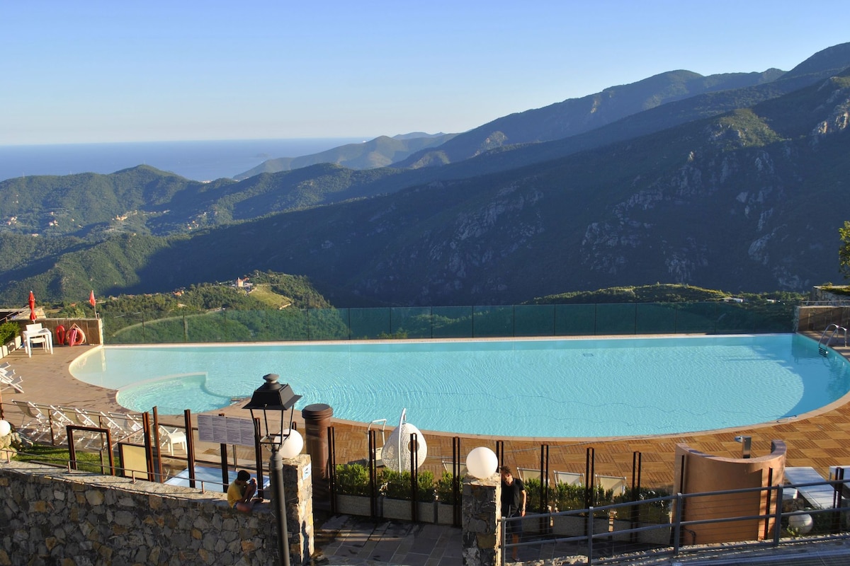 An infinity pool is seen overlooking rolling green hills and distant mountains. The water appears calm, reflecting the clear blue sky above. Sun loungers are arranged around the pool area, and the ocean is visible in the background.