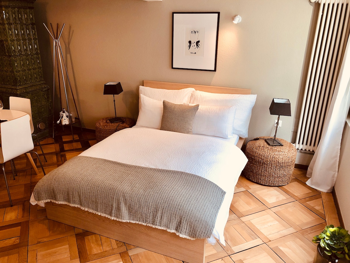 A cozy bedroom is furnished with a spacious bed, complemented by soft bedding and decorative pillows. Two bedside lamps provide gentle illumination, while natural light enters from the nearby window. A small table and seating are visible, enhancing the comfortable atmosphere.