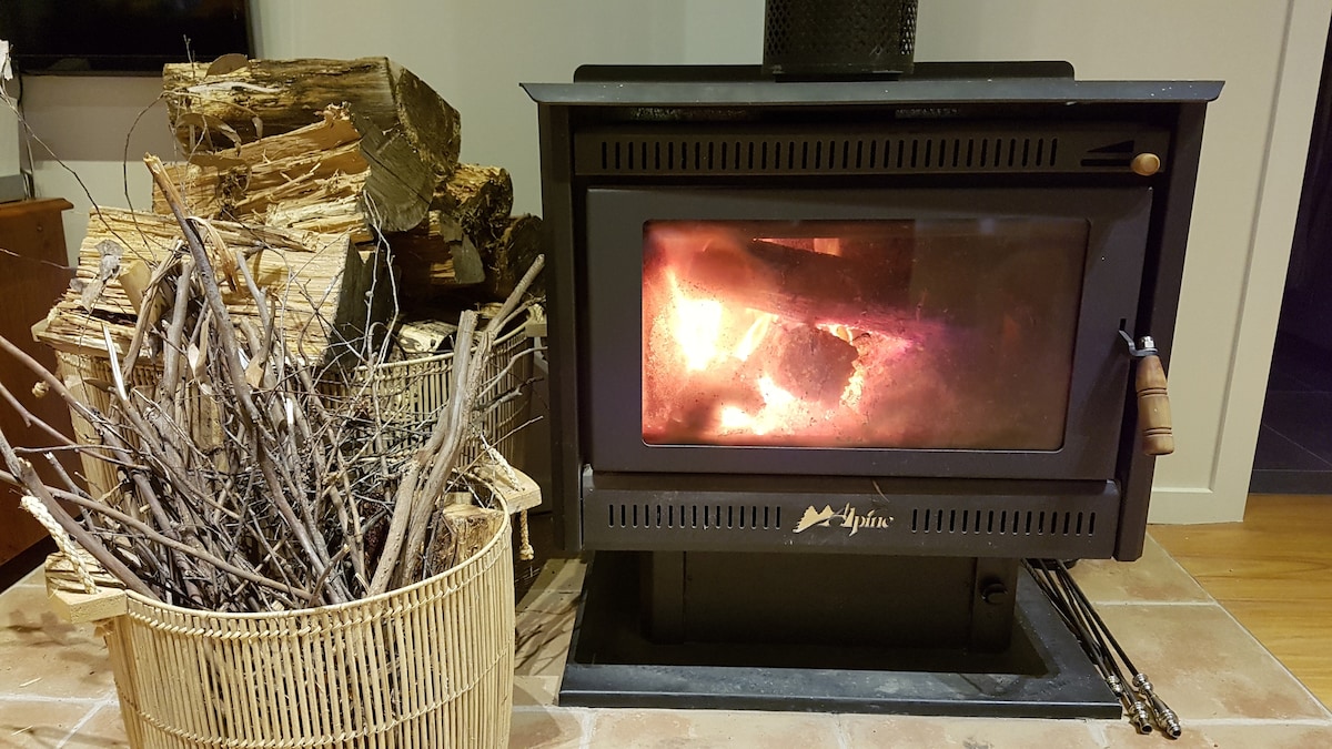 A wood-burning stove is positioned against a tiled floor, its glass door revealing warm flames inside. Beside the stove, a basket filled with firewood and kindling is neatly arranged, adding to the cozy ambiance of the living space.