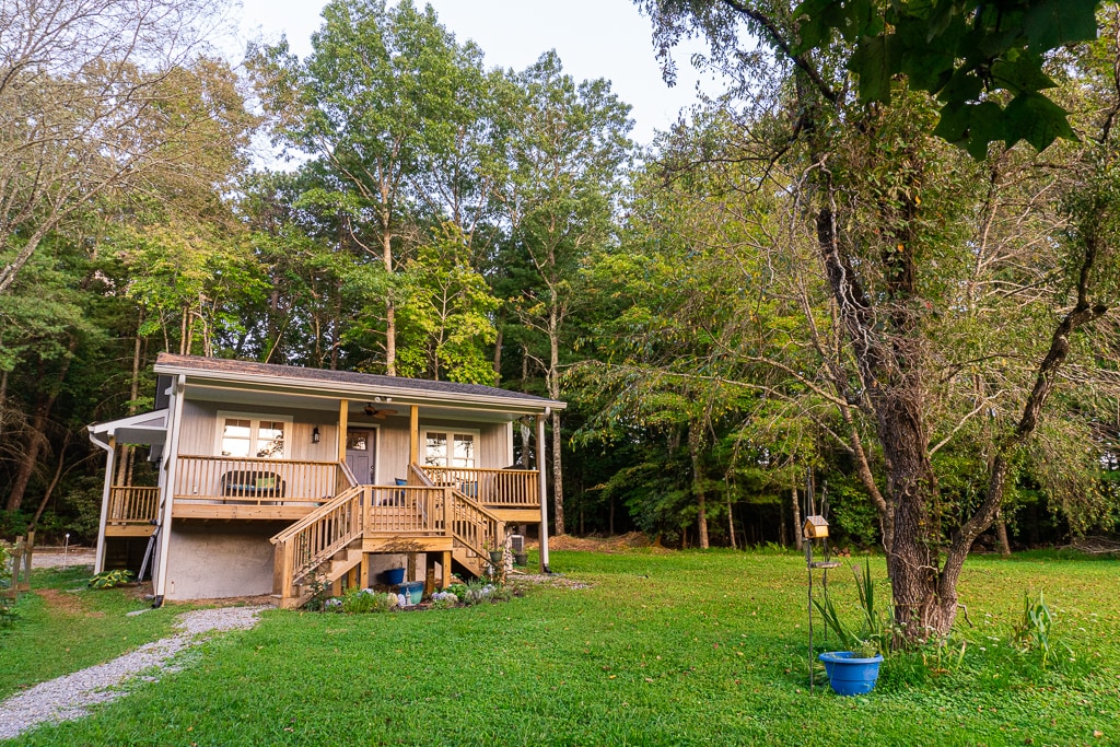 A modern cottage is set within a lush green yard, surrounded by trees. The property features a welcoming porch with a wooden railing and steps leading up to the entrance. A small gravel path is visible in the foreground, enhancing the inviting outdoor space.