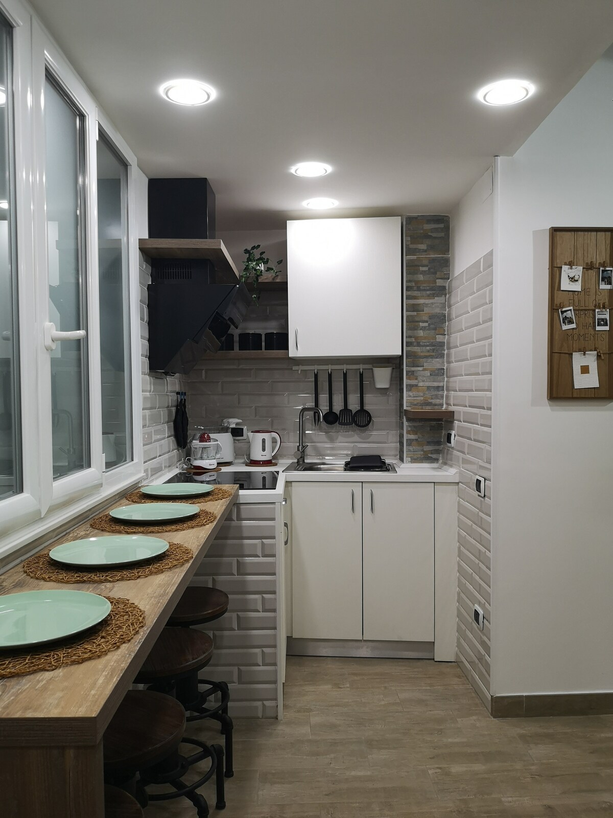 A modern kitchen space is shown, featuring a compact layout with white cabinetry and grey tiled walls. Countertop appliances like a kettle are visible, along with cooking utensils neatly arranged on the wall. Four round placemats adorn the wooden bar, with large windows allowing natural light.