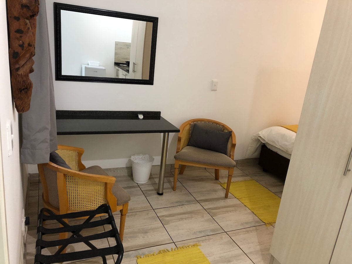 A cozy corner of the self-catering unit features a wooden table with two chairs. A mirror adorns the wall above the table. The floor is tiled with light-colored tiles, and a small wastebasket sits near the table. A spacious bed is visible in the background.