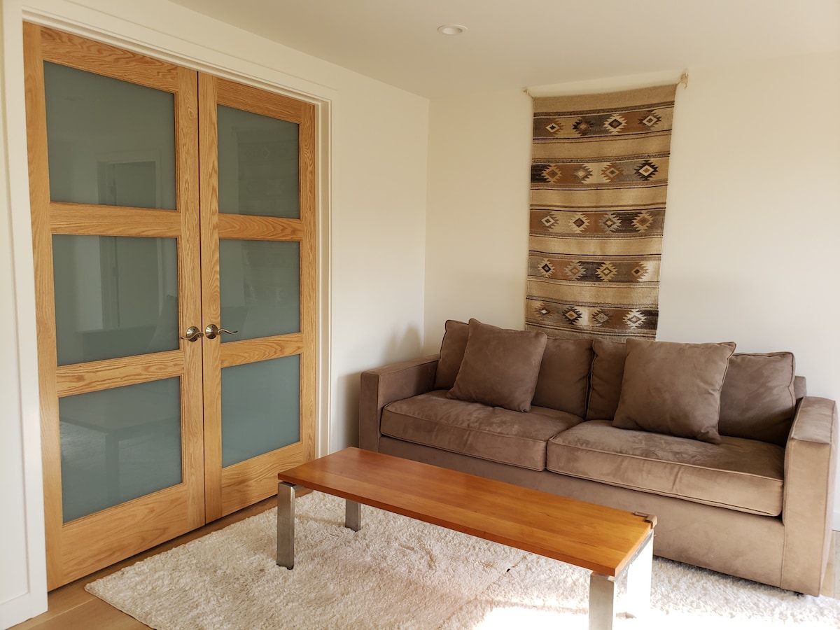 A cozy sitting area features a brown sofa complemented by a wooden coffee table. Light-colored walls enhance the space, while a decorative wall hanging adds visual interest. Double doors with frosted glass panels provide an inviting transition to another room.