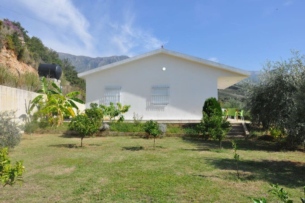 A simple white house is set against a backdrop of rolling hills, surrounded by a lush garden. Various small trees and shrubs dot the green lawn, providing a serene outdoor space. The clear blue sky complements the overall tranquil setting.