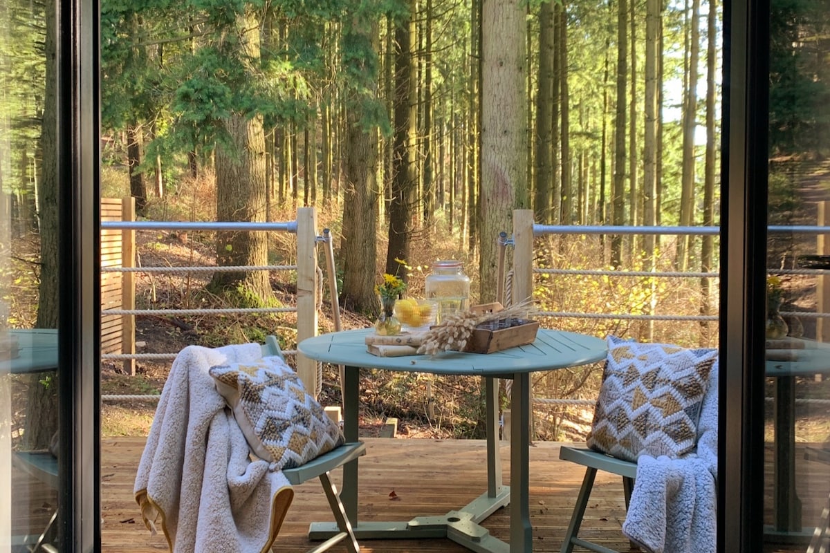 A round wooden table with two patterned chairs is set on a terrace, surrounded by tall trees. A cozy blanket drapes over one chair, while a tray with refreshments rests on the table, inviting enjoyment of the tranquil forest views.