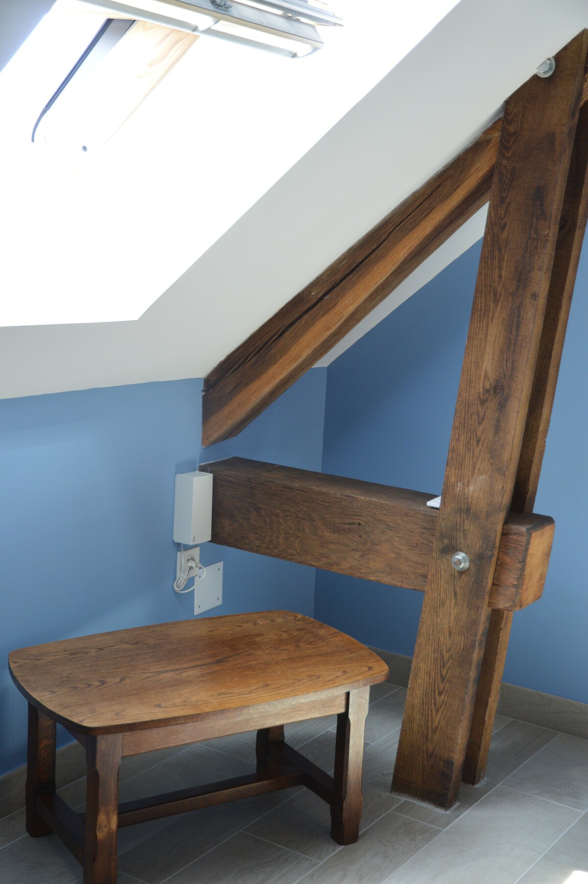 A light-filled corner features a wooden stool positioned against a wall with exposed beams. A skylight above provides natural illumination, highlighting the blue wall. The wooden structure complements the tiled floor, creating a cozy nook within the room.