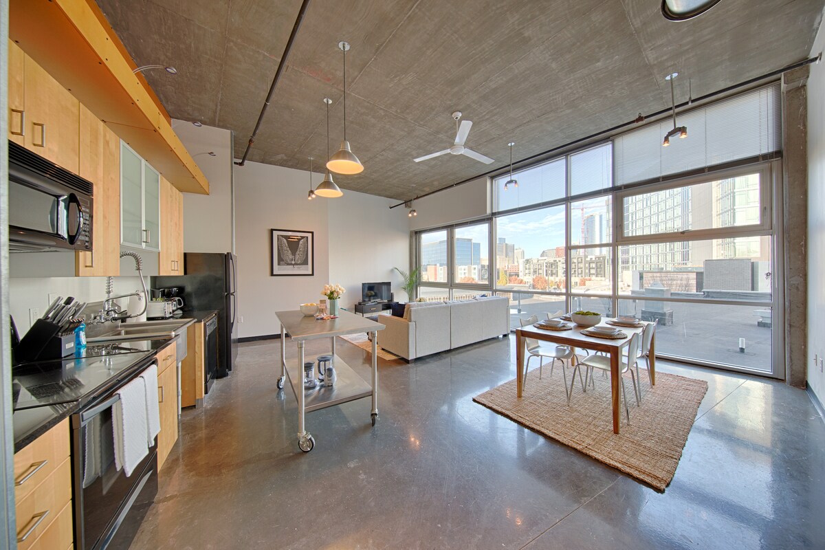 A modern open-concept living space is presented, showcasing a fully equipped kitchen with wooden cabinetry and stainless steel appliances. A dining table with seating for four is located next to the kitchen, while natural light floods in through large windows, illuminating the spacious living area with a comfortable sofa and stylish decor.