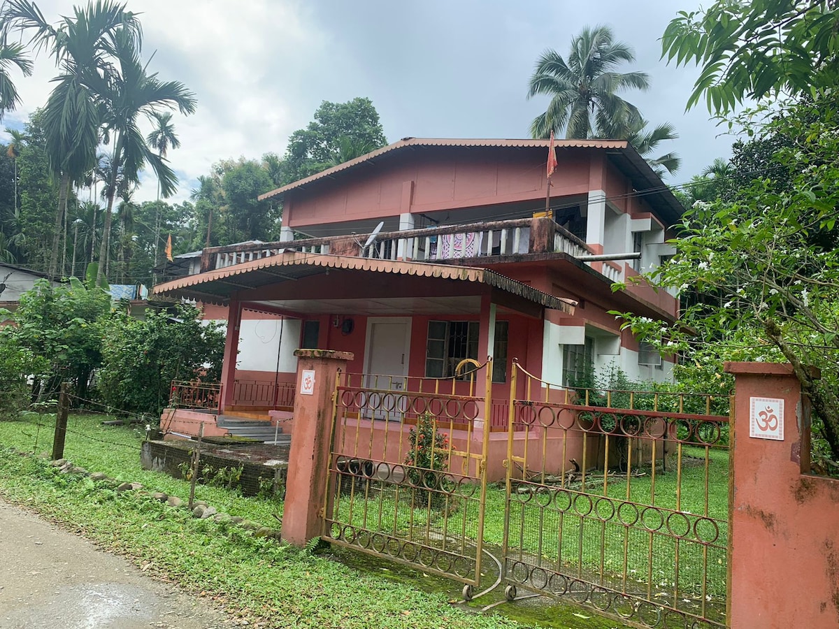 A two-story bungalow with a relaxed design is set among lush greenery. The exterior features a reddish façade with a covered terrace, surrounded by a decorative metal fence. Tall palm trees offer shade, while colorful laundry hangs from the balcony.