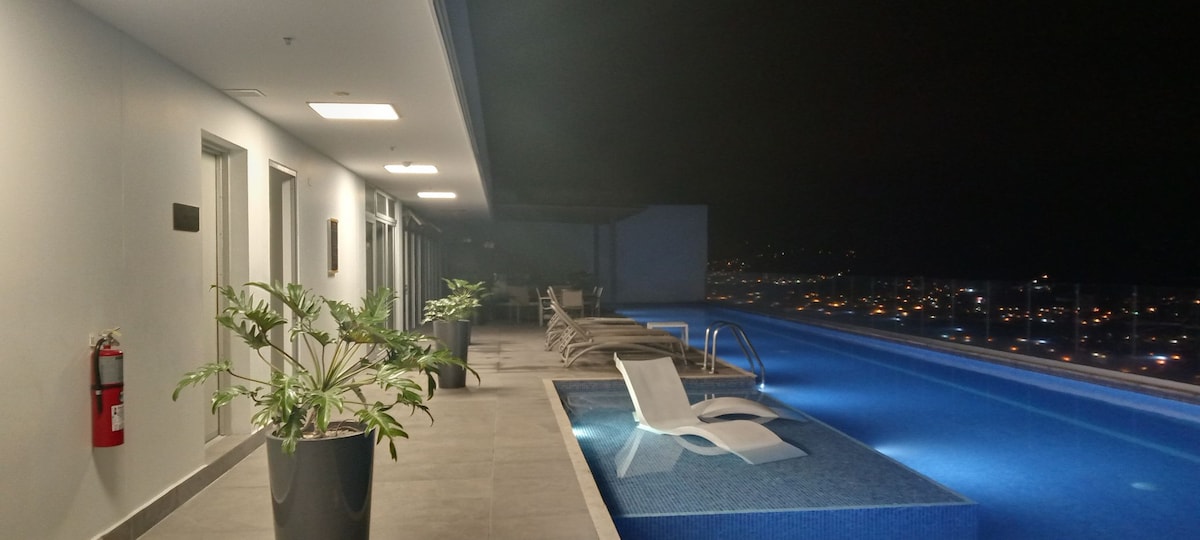 An illuminated rooftop area features a heated pool overlooking the city, with lounge chairs positioned along the edge. Potted plants add a touch of greenery, while the calm water reflects the evening lights, enhancing the serene ambiance of the space.