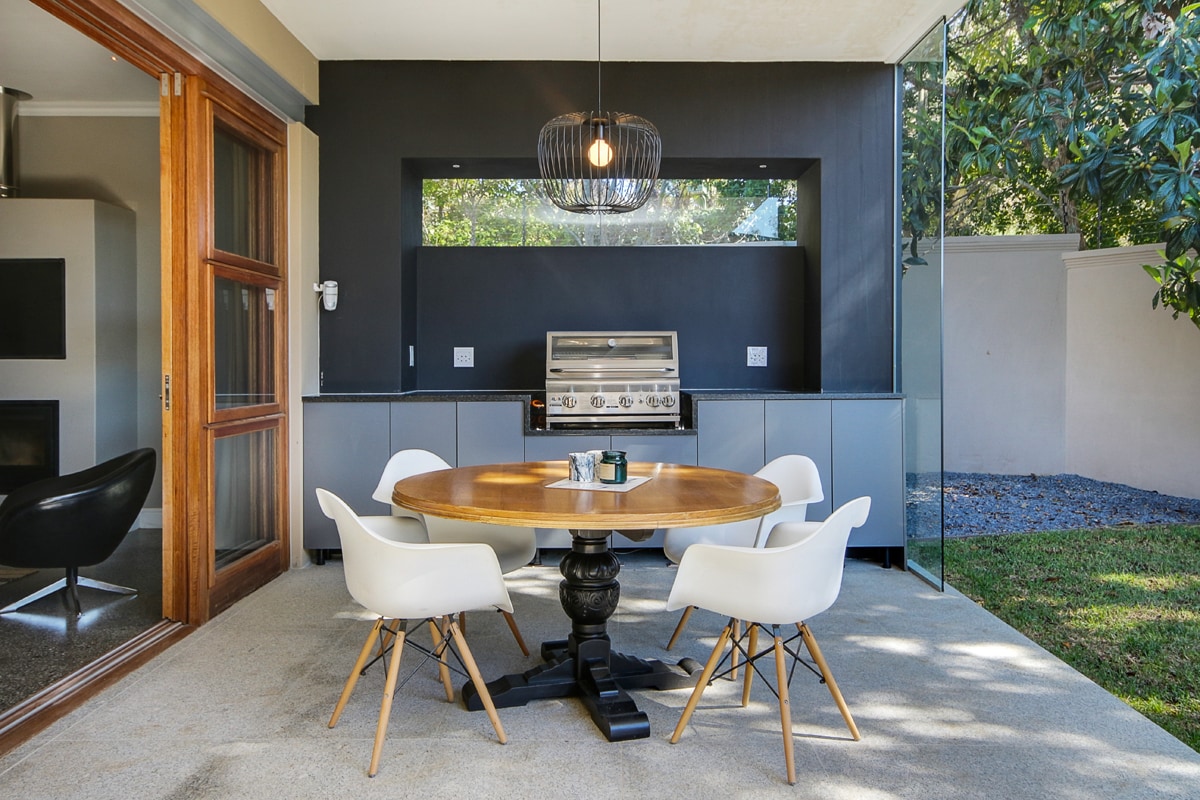 A modern outdoor dining area features a round wooden table surrounded by four white chairs. A sleek BBQ unit is positioned against the dark wall, complementing the natural light that enters through the large glass panels. Lush greenery is visible in the background.