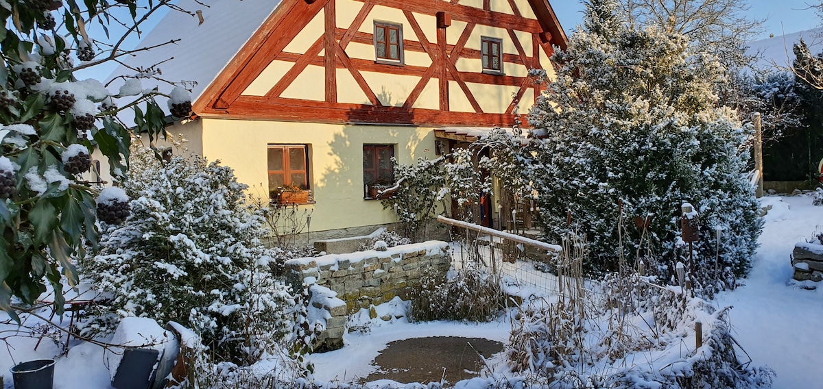 A traditional half-timbered house is surrounded by a winter landscape covered in snow. Thorny shrubs and greenery are visible in the foreground, while the house features a warm yellow exterior and wooden accents. Frosted trees line the property, creating a serene atmosphere.