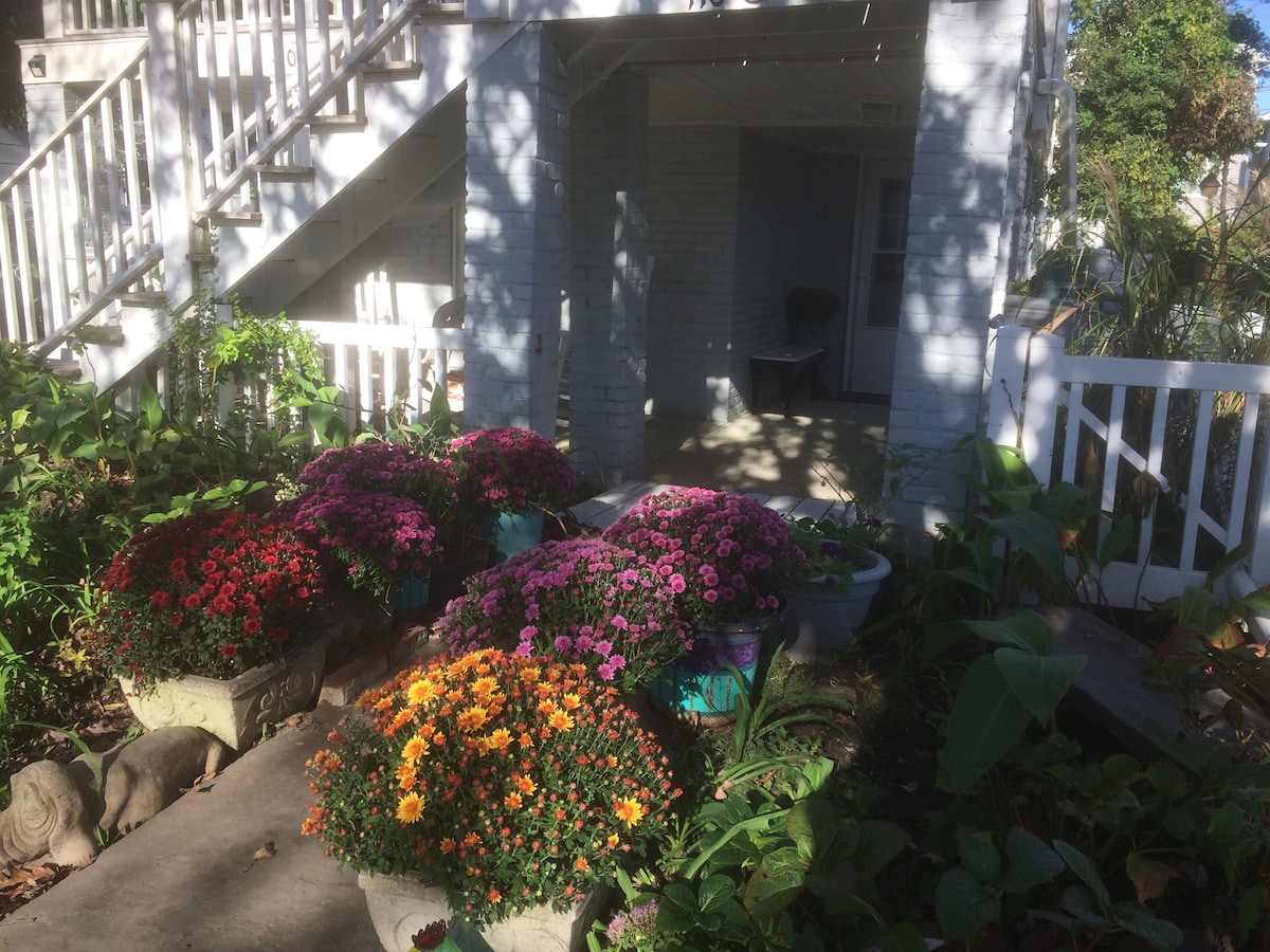 Brightly colored flower pots display a variety of blooming flowers, including purple, red, and orange hues. The entrance is framed by lush greenery and a white railing, with a glimpse of a light-filled patio area visible through the doorway.