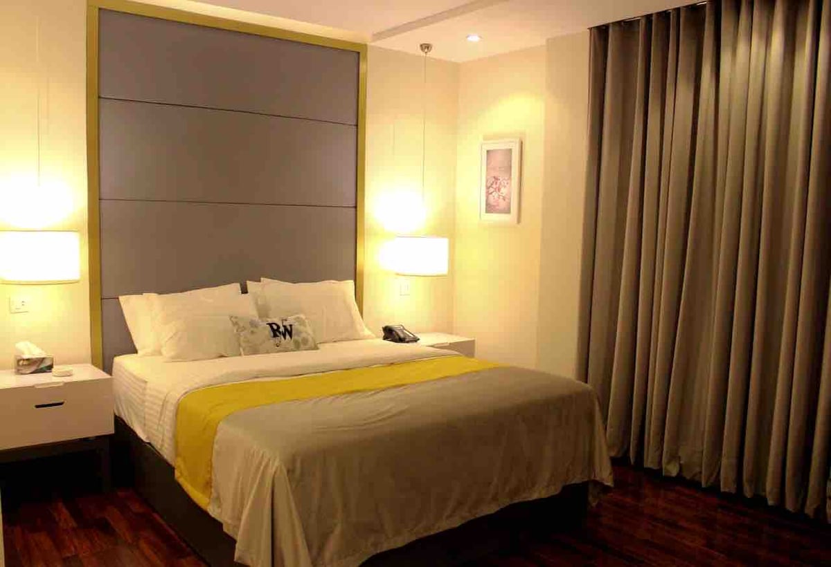 A modern room features a spacious bed with a light-colored bedspread and a bright yellow accent. Soft lighting is provided by bedside lamps on either side. Elegant curtains frame a window, complementing the minimalist decor.