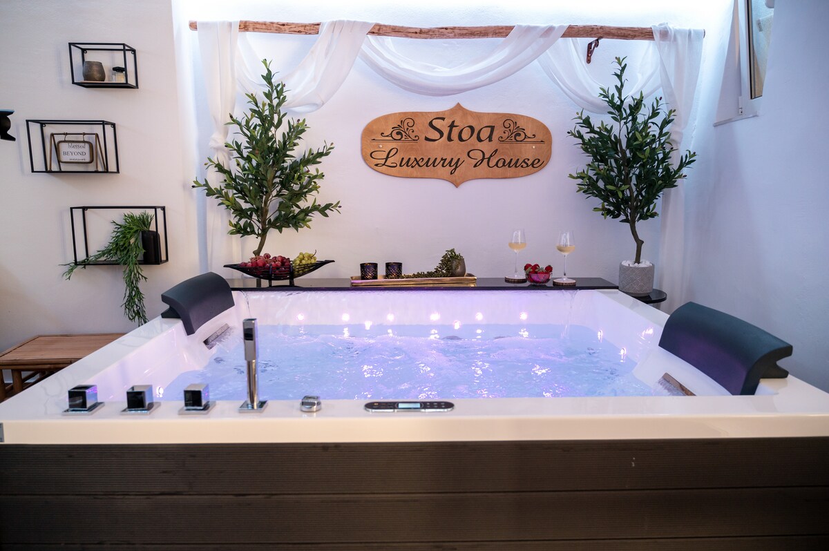 A relaxation room features a heated spa whirlpool surrounded by greenery, enhancing the tranquil setting. A wooden sign displaying 'Stoa Luxury House' is mounted on the wall. Seated areas, decorative fruits, and wine glasses complete the inviting atmosphere.