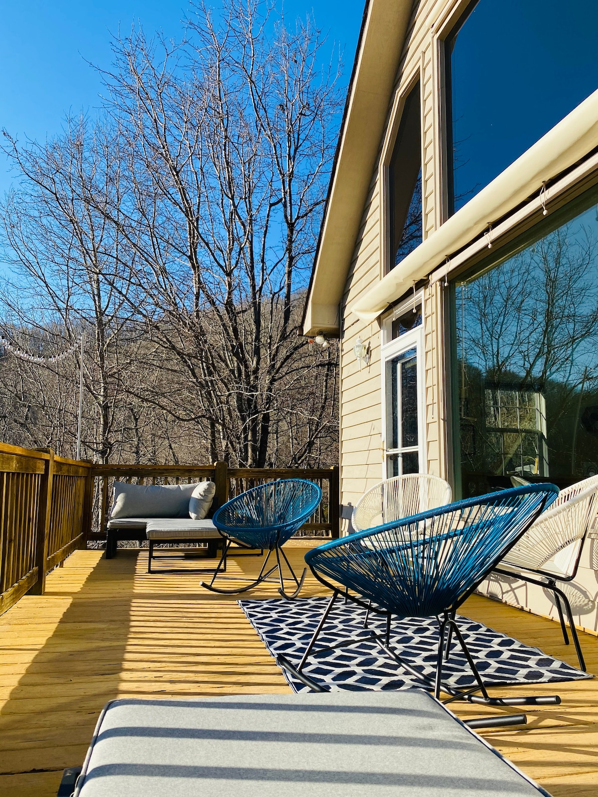 A spacious wooden deck is furnished with several lounge chairs and a comfortable seating area. A patterned rug adds visual interest, while large windows provide expansive views of the surrounding nature. Leafless trees in the background indicate a seasonal transition.