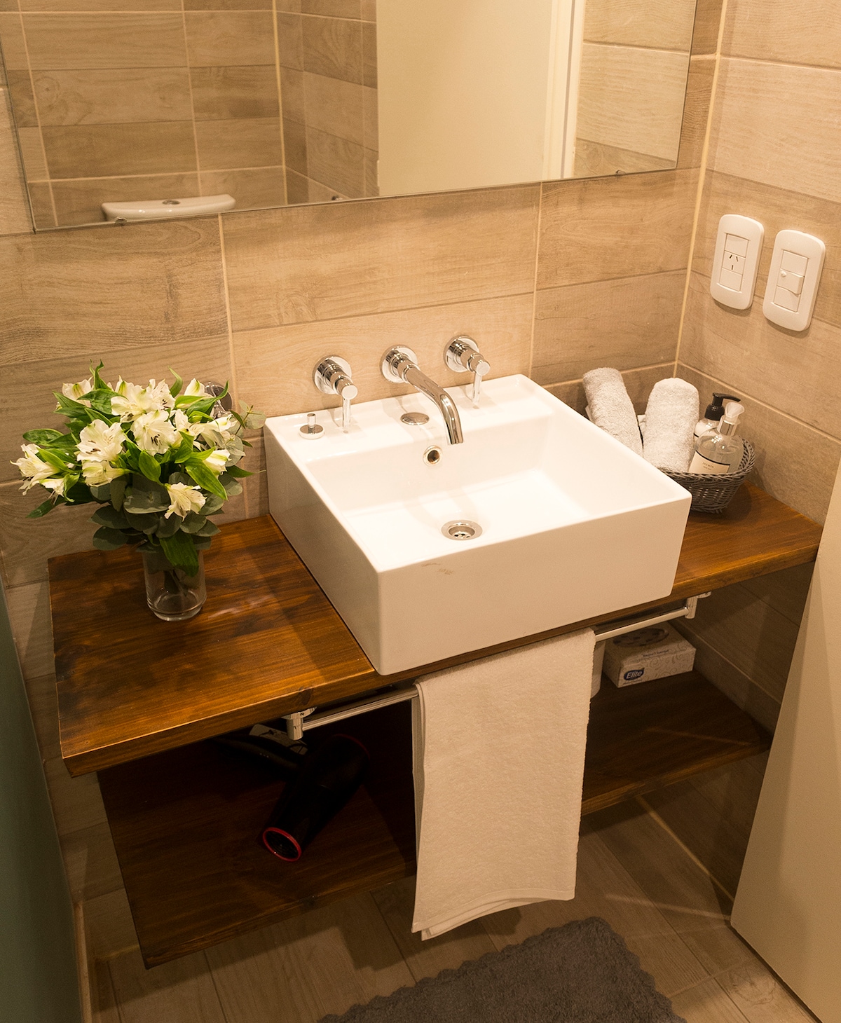 A modern bathroom sink is positioned atop a wooden shelf, accompanied by a small vase of white flowers. Rolled towels and a hairdryer are arranged neatly beside the sink, while a mirror reflects light and enhances the space.