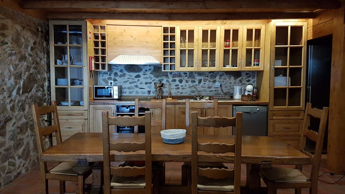 A spacious kitchen is presented, featuring wooden cabinetry with glass-fronted shelves. A large wooden dining table is centered in the room, surrounded by several chairs. Modern appliances including a microwave and dishwasher are visible, and a stone accent wall adds texture.