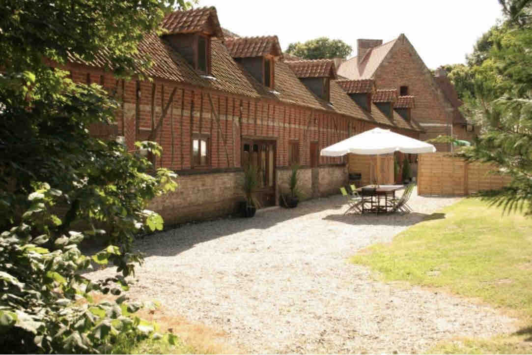 The exterior of a historic cottage complex is depicted, showcasing traditional wooden beams and brickwork. An outdoor dining area with a table and chairs is positioned beneath a large umbrella, surrounded by a gravel driveway and open green lawn, leading to landscaped gardens.