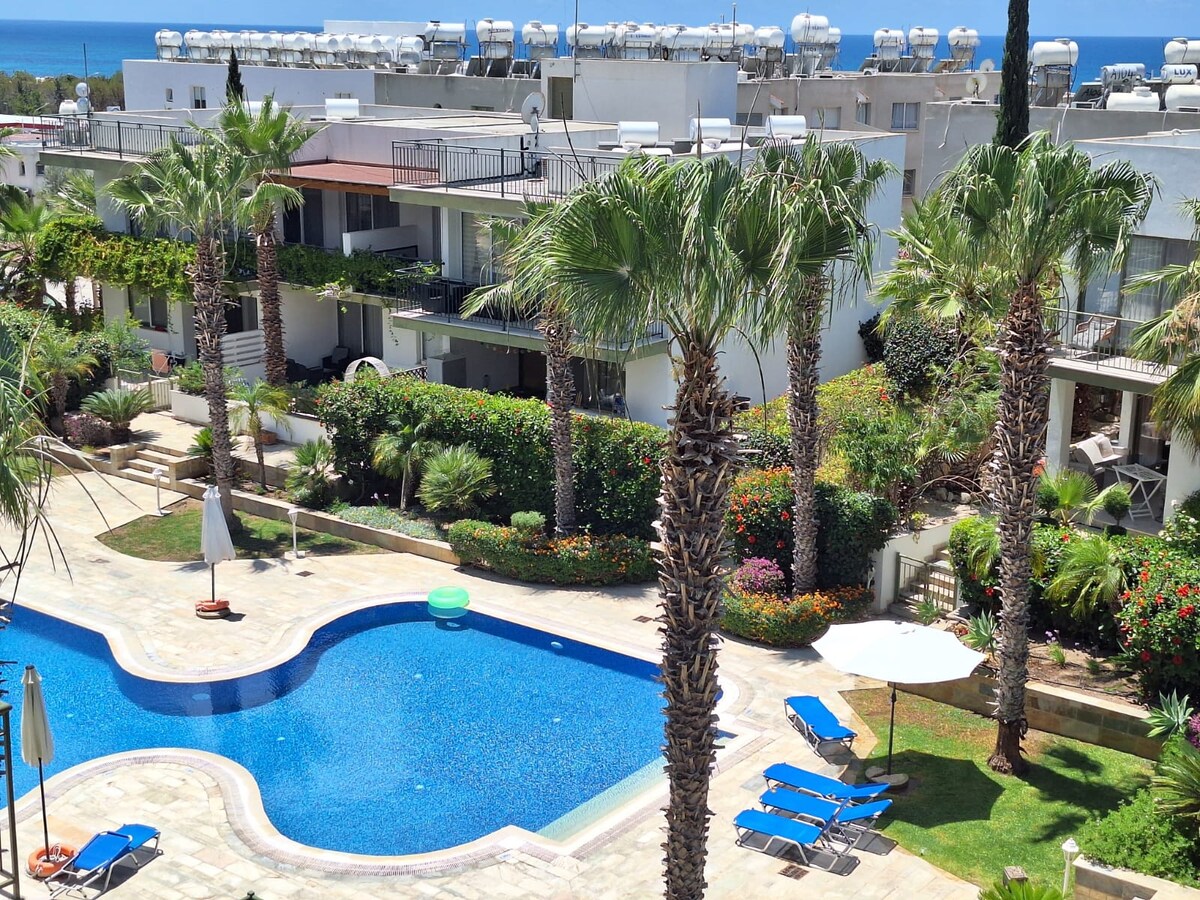 A well-maintained outdoor area features a communal swimming pool with clear blue water surrounded by lounge chairs. Lush palm trees and vibrant landscaping enhance the space. Sun loungers and umbrellas are arranged for guest comfort, with buildings visible in the background.