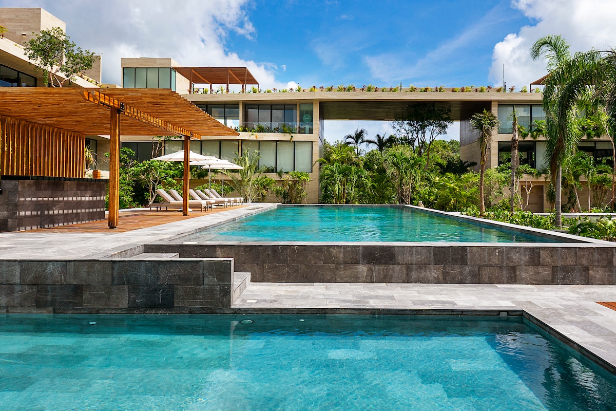 The image showcases two outdoor pools framed by lush greenery and modern architecture. Numerous sunbeds with cushions are visible around the pool area. A wooden pergola provides shade near the pool, enhancing the serene environment.