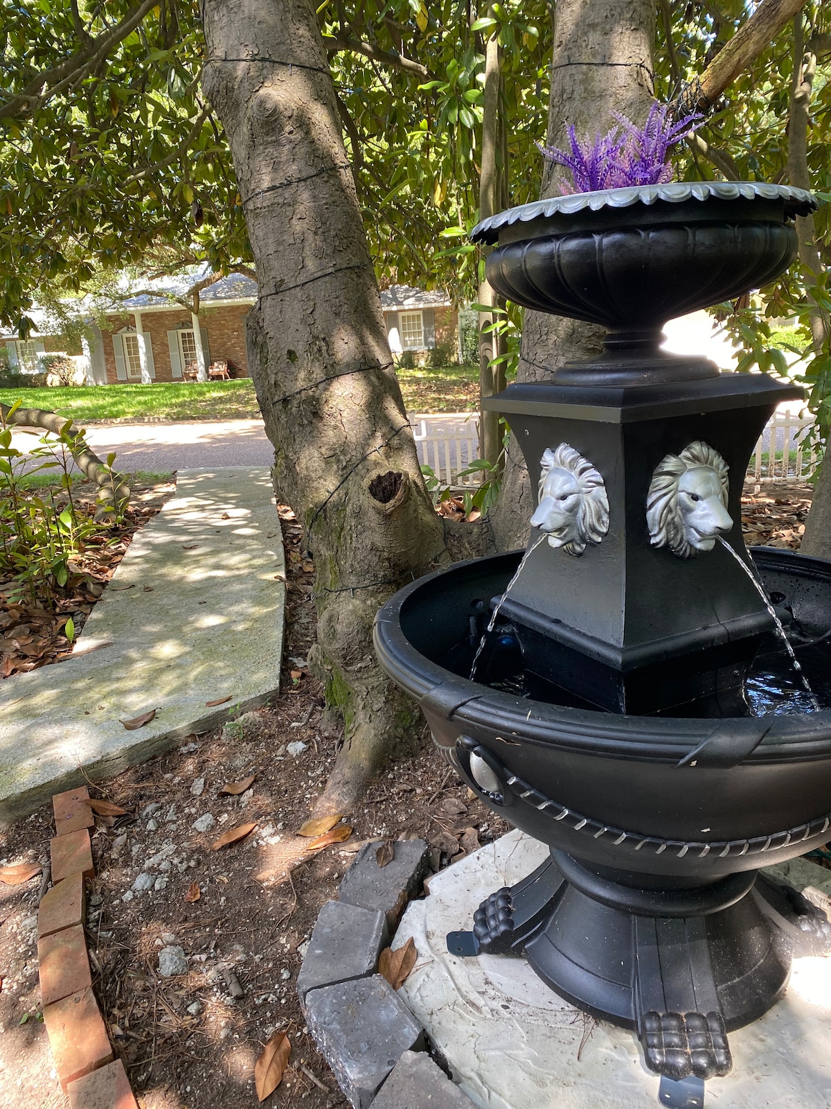 A decorative fountain is positioned in a garden, featuring lion-head spouts with water flowing gently. The fountain is surrounded by textured soil and bricks, with a small cluster of purple flowers placed on top. Tall magnolia trees provide a natural backdrop.