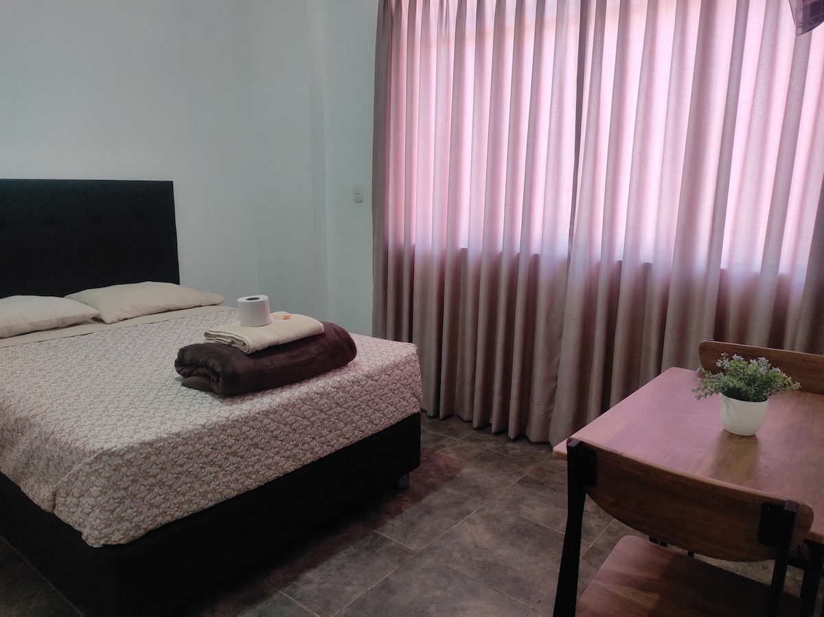 A comfortable room is showcased, featuring a spacious bed with soft linens and pillows. Natural light filters through the sheer curtains, creating a calming atmosphere. A wooden table with a small plant adds a touch of greenery to the space.