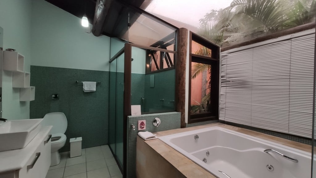 A modern bathroom features a spacious jetted tub beside a large window with natural light. A walk-in shower is enclosed in glass, and the walls are accented with green tiles. Additional natural elements are visible through the window, enhancing the serene environment.