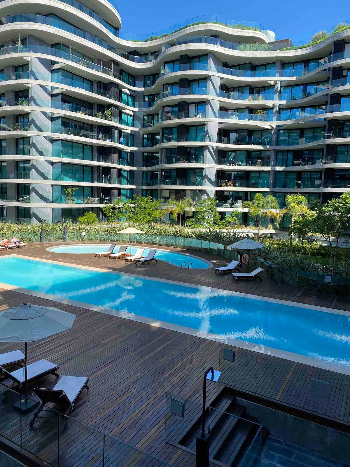 A spacious outdoor area features a large swimming pool surrounded by a wooden deck. Sun loungers are positioned nearby under umbrellas. Lush greenery and modern architecture create a serene environment, showcasing the building's glass facades reflecting natural light.