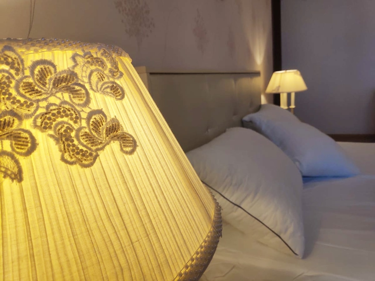 A well-lit sleeping area features a spacious bed with soft white linens and two pillows. An elegant bedside lamp with a detailed shade casts a warm glow, complementing a second lamp positioned in the background.